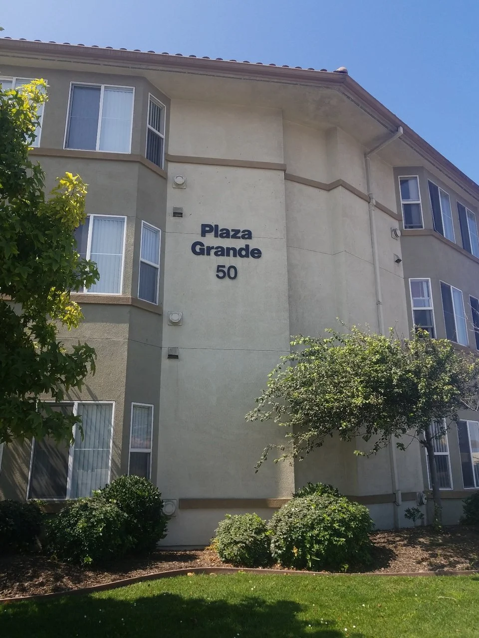 Apartment building with a sign that reads 'Plaza Grande 50' and features multiple windows, trees, and bushes outside.