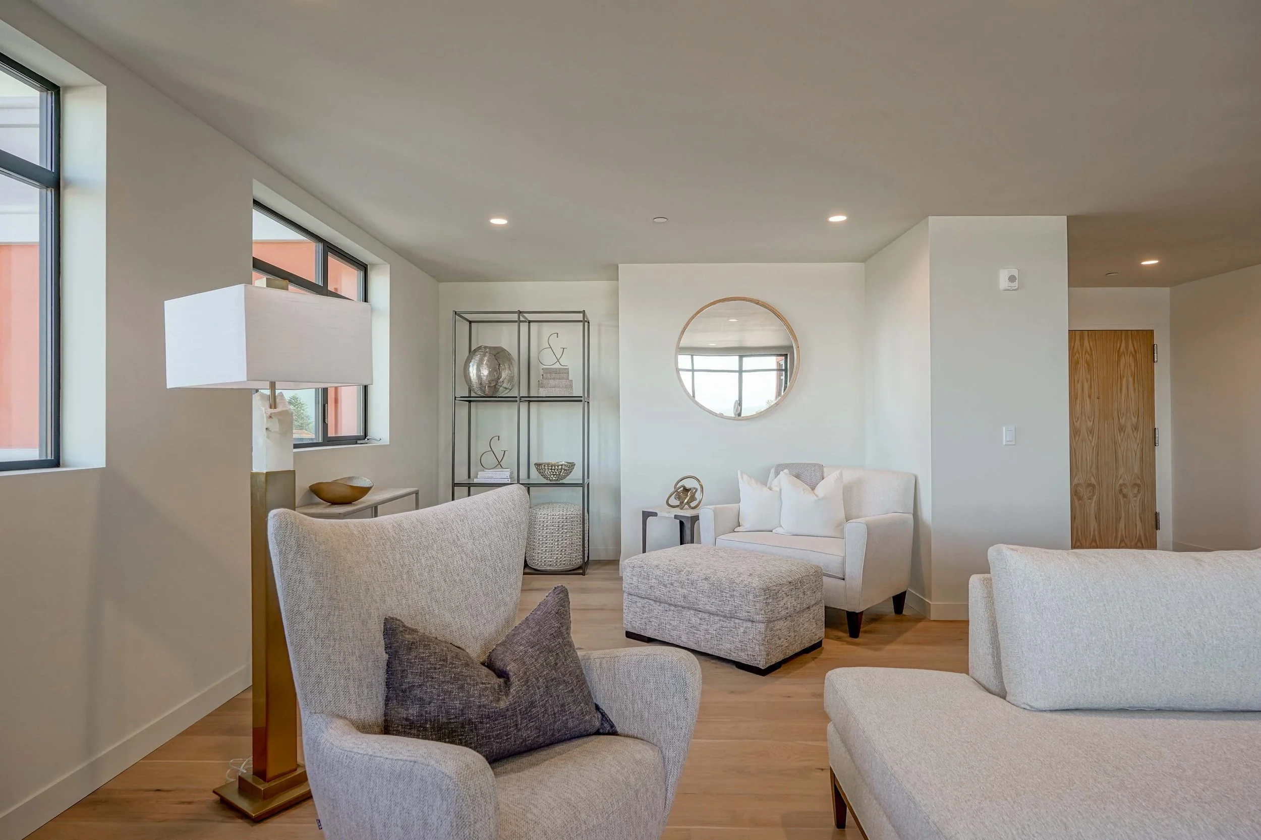 Bright living room with beige chairs, a sofa with white pillows, a round mirror on the wall, windows, shelves, and wooden flooring.