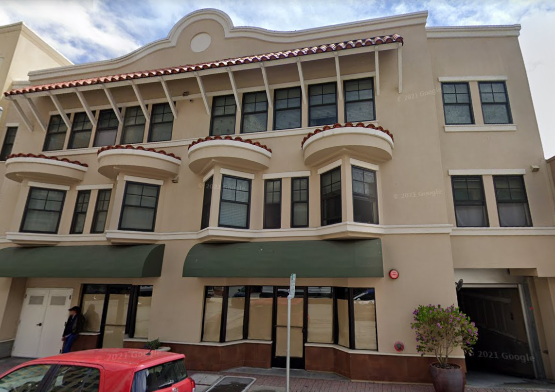 Multi-story beige building with four bay windows on the second and third floors, red-tiled awnings, and a green awning covering the ground-floor windows and entrance. There is a parked red car and a person standing near the entrance on a sidewalk wit