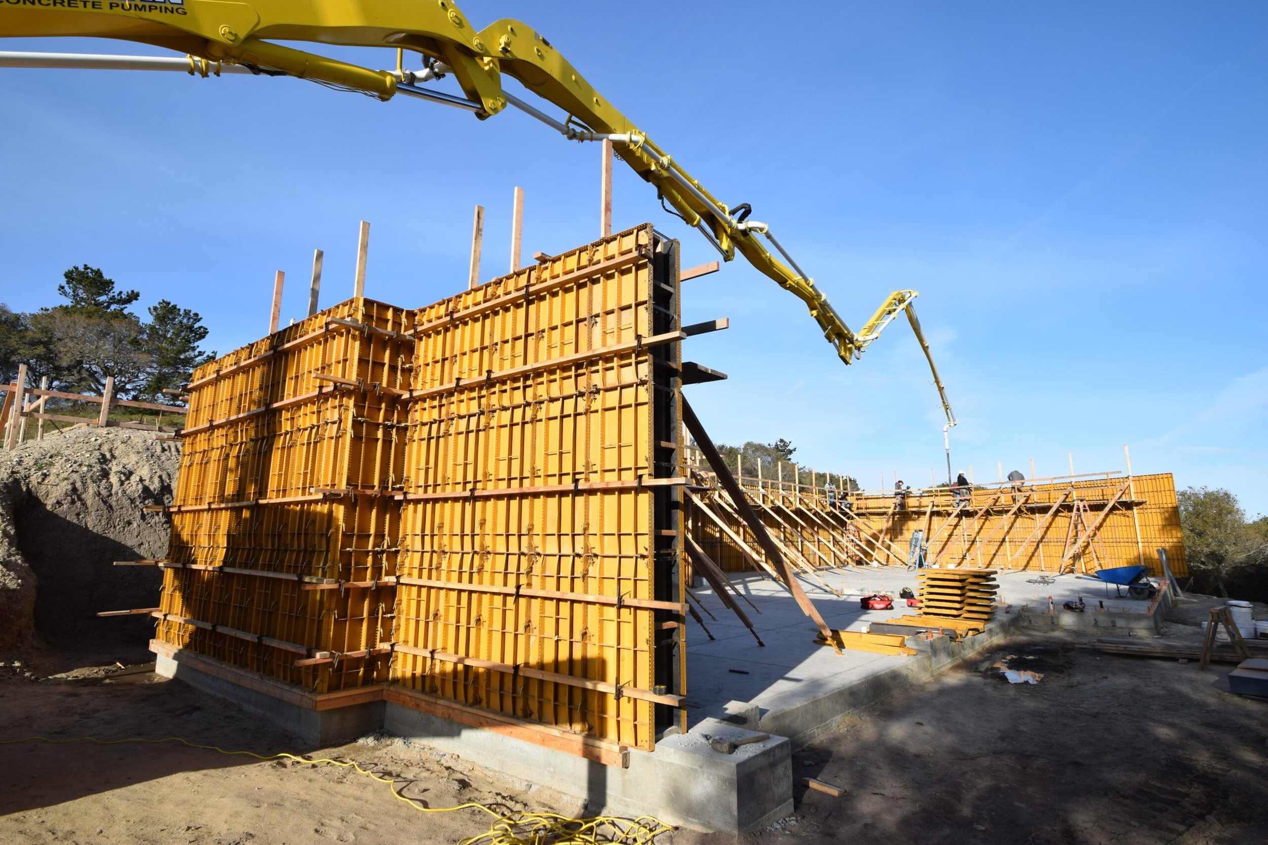 Construction site showing concrete wall forms in yellow with stairs leading up, a concrete foundation, and a large yellow concrete pump extending over the site against a clear blue sky.