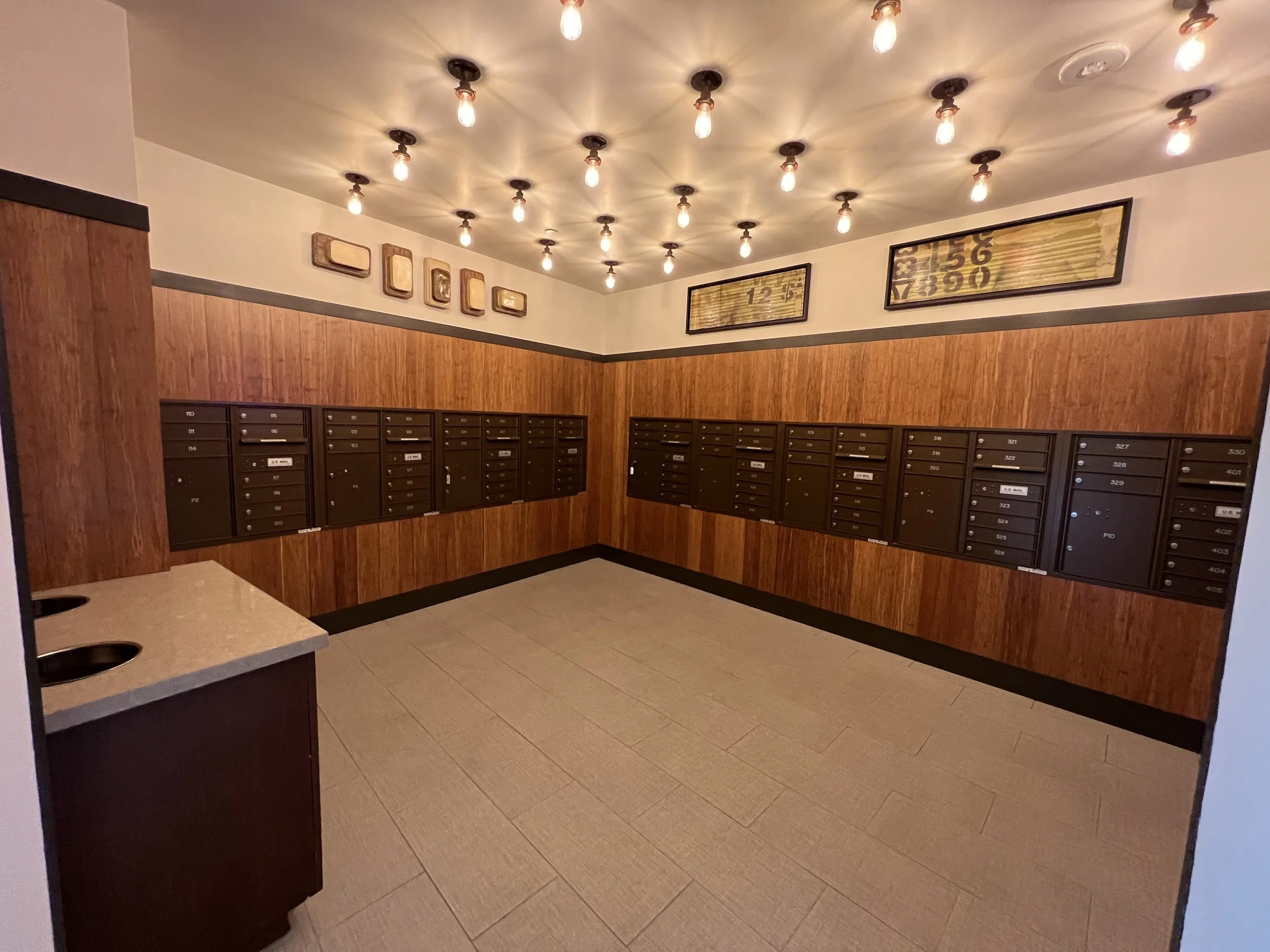 Hallway with black mailboxes lining the wall, framed artwork, warm ceiling lights, and beige tile flooring.
