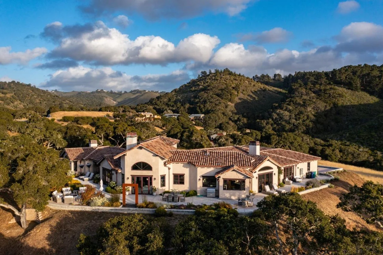 Luxury house with a terracotta tiled roof on hillside surrounded by green trees and rolling hills under a partly cloudy sky.
