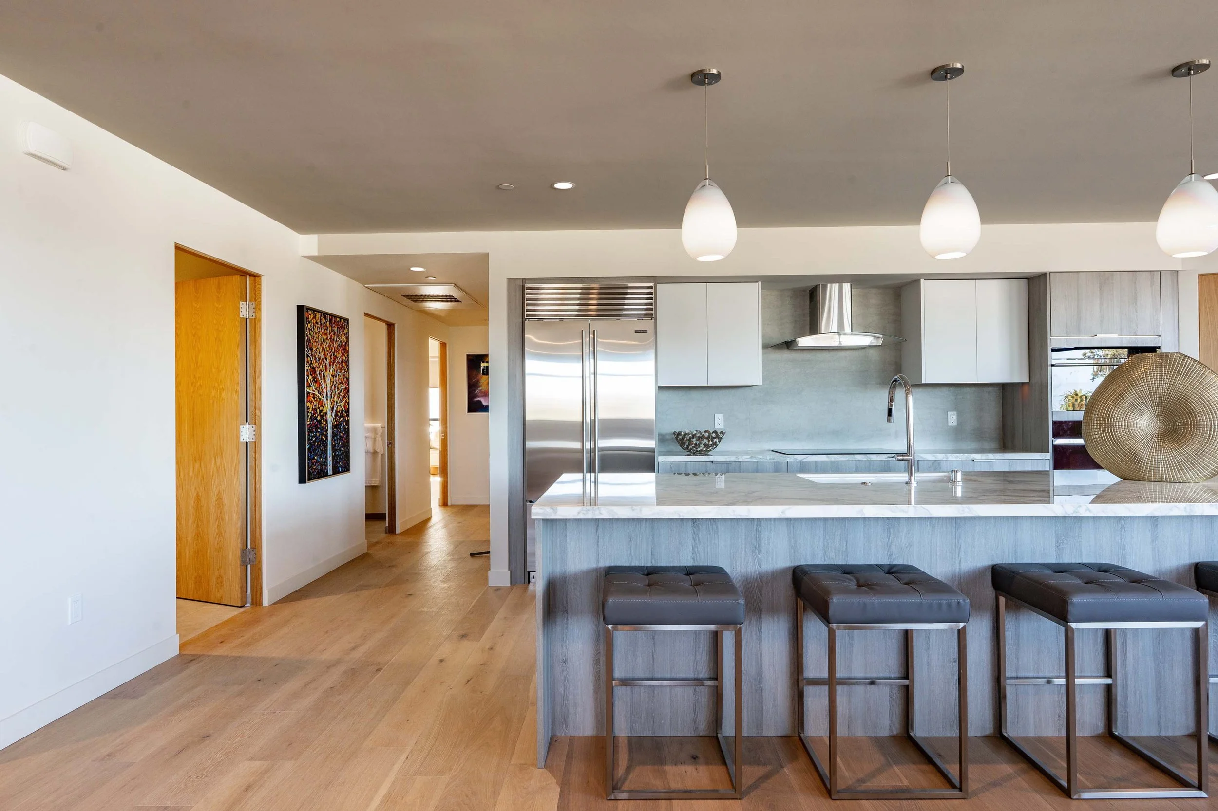 Modern kitchen with white cabinetry, stainless steel refrigerator, marble countertops, three pendant lights hanging from the ceiling, and wooden floors. There is a seating area with dark leather stools at the island.