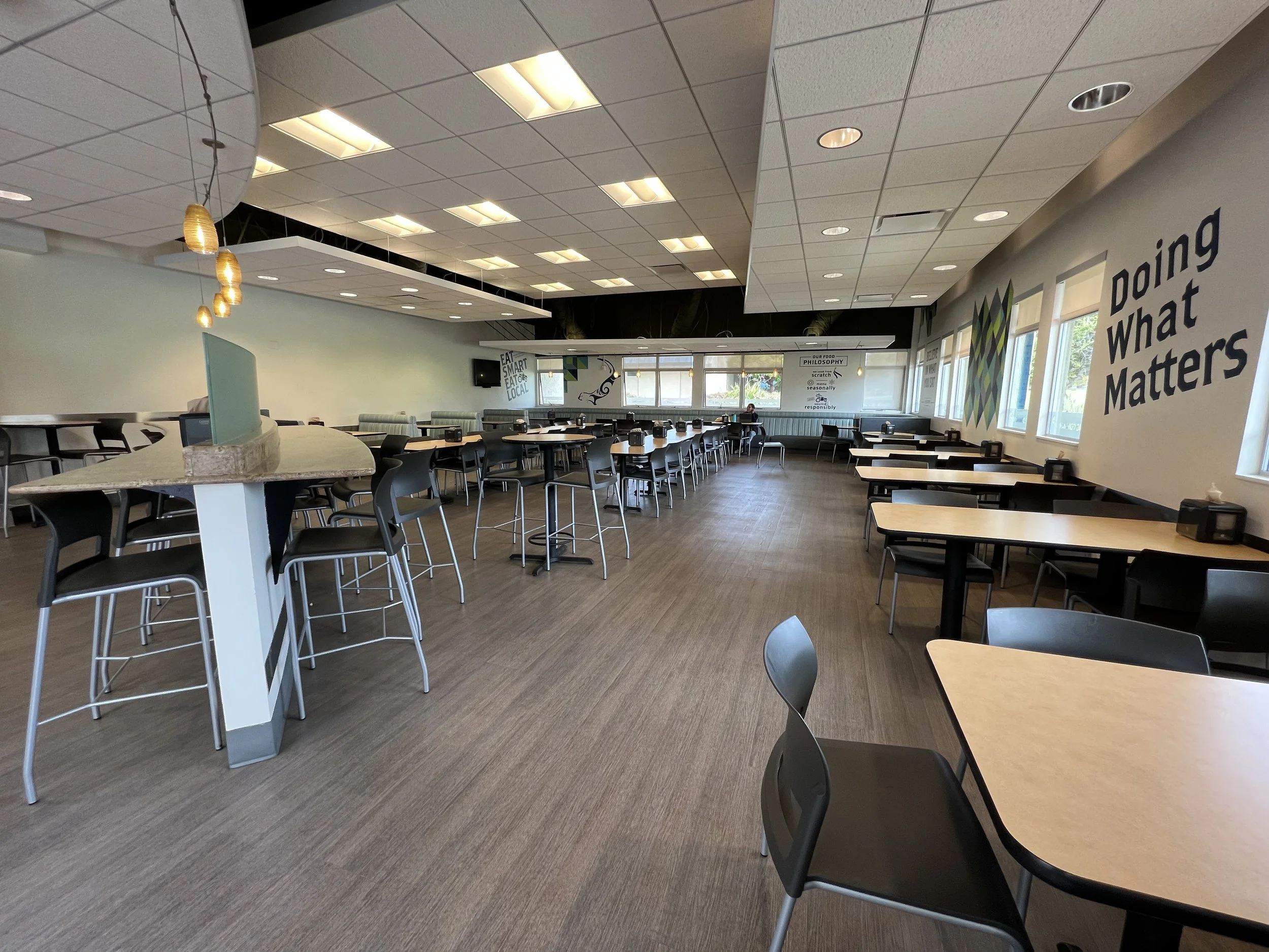 Empty dining area with several tables and chairs arranged neatly, windows with blinds on the side, and motivational posters on the walls, including one that says "Doing What Matters".