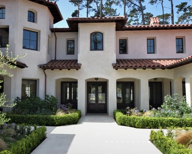 Front view of a Mediterranean-style house with white stucco walls, a red tiled roof, and large windows. The entrance has three black doors, and lush green landscaping with bushes and flowers surrounds the walkway leading to the front door.