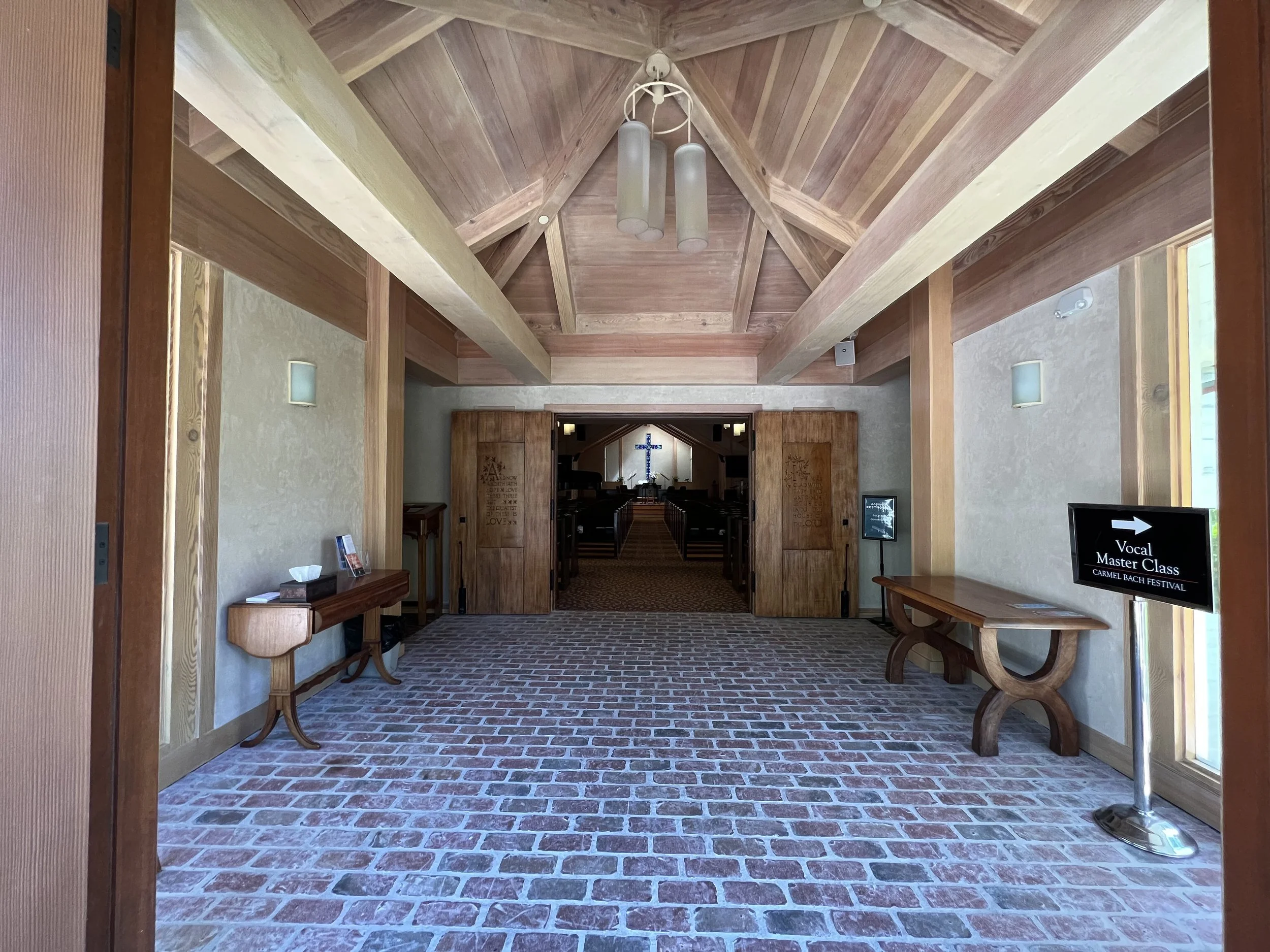 Interior view of a church hallway with wooden beamed ceiling, brick floor, and pews leading to an altar with a cross at the back, illuminated by natural light.