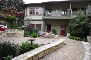 A patio area in a backyard with pavers, a curved stone wall, a bird bath, and outdoor furniture in front of a two-story house with a balcony.