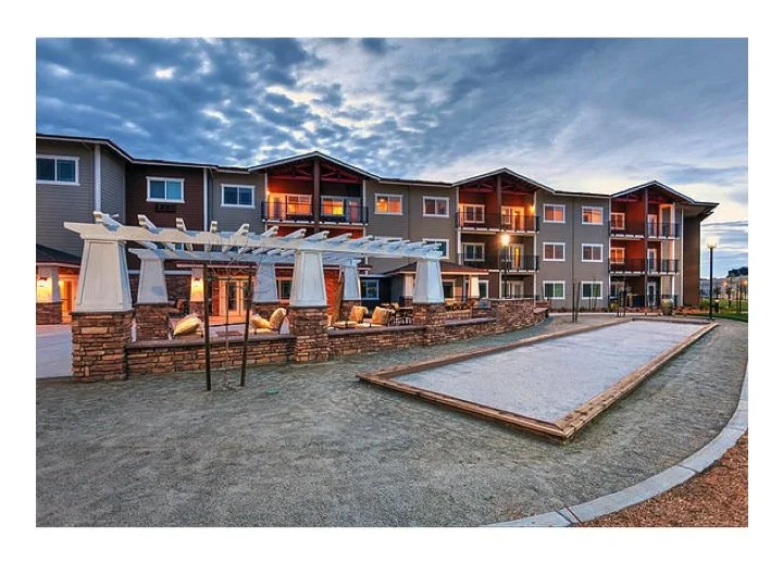 Apartment complex with outdoor courtyard, sand volleyball court, and a shaded seating area with white pillars, during early evening with partly cloudy sky.