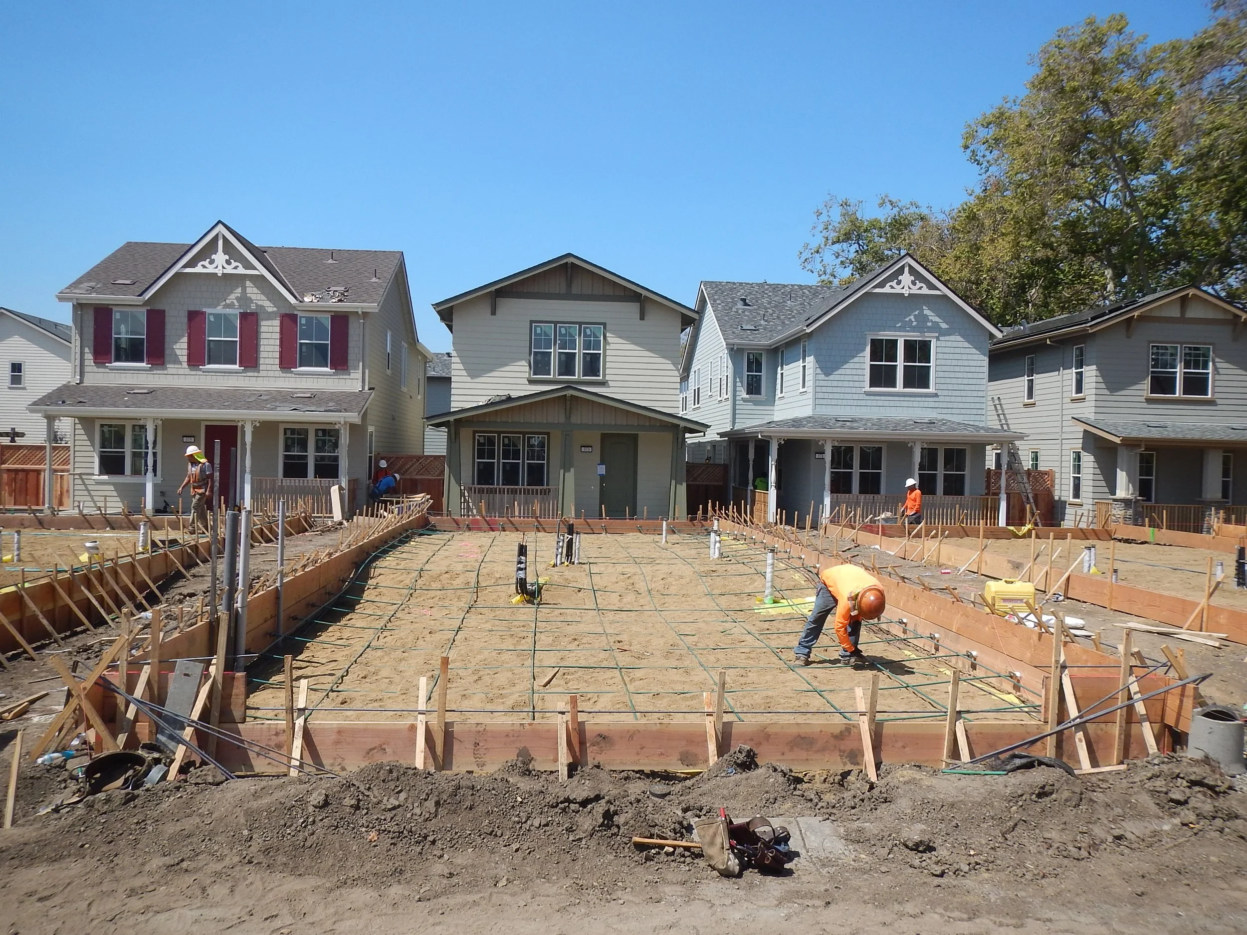 Construction site with workers building a house, wooden frameworks, and residential houses in the background.