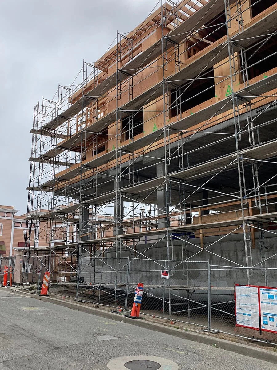 Construction site with scaffolding around a multi-story building in progress, showing wooden framing and concrete supports, with safety cones and fences at street level.