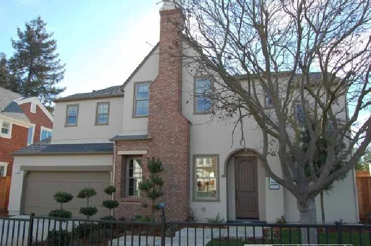 Front view of a two-story house with a brick chimney, beige stucco exterior, and a brown front door, surrounded by a metal fence and landscaped with small trees and a large leafless tree.
