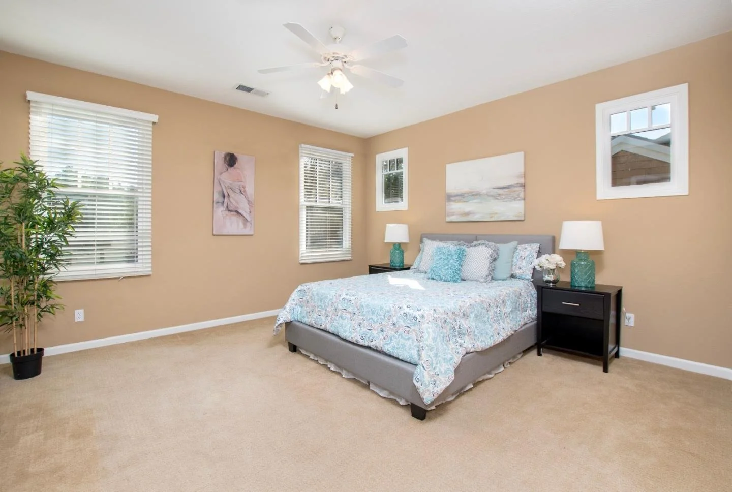 A bedroom with beige walls, a ceiling fan, three windows with blinds, a bed with light blue bedding, two black nightstands with lamps, a potted plant, and wall art.