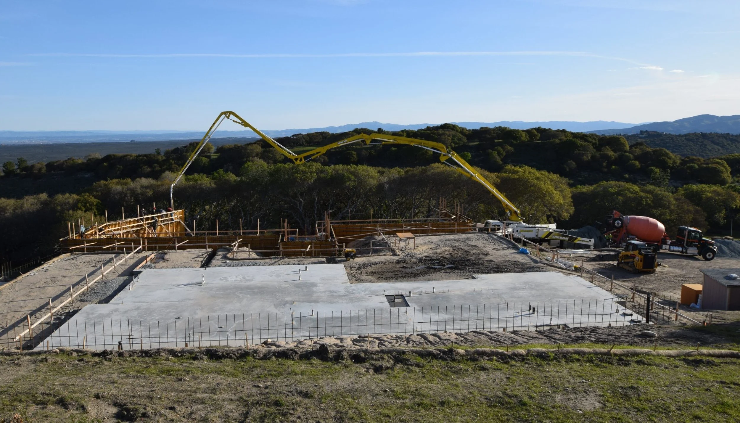 Construction site on a hill with concrete foundation, wooden framing, and a large yellow cement pump truck pouring concrete, surrounded by green trees and distant mountains.