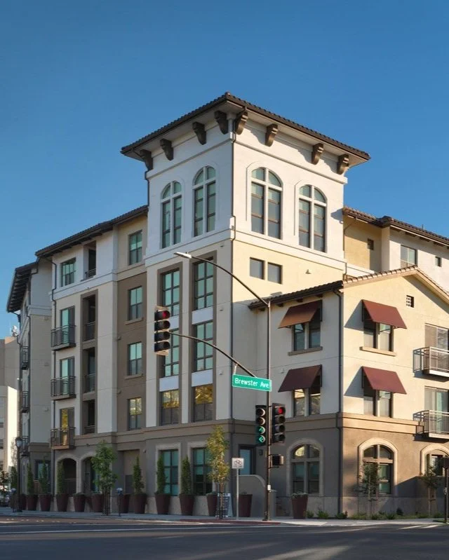 A multi-story residential building with a Spanish architectural style, featuring arched windows and balconies, located on the corner of Brewster Avenue, with traffic lights and potted plants in front.