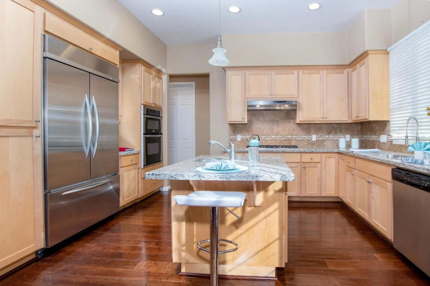A kitchen with wooden cabinets, a granite countertop island, stainless steel appliances, a double-door refrigerator, a double oven, a gas stove with stainless steel range hood, a dishwasher, and a large window with blinds, hardwood flooring, and a ha