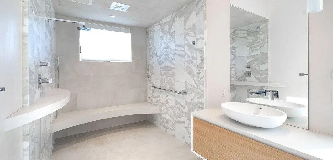 Modern bathroom with marble walls and flooring, a built-in bench, a window, and two vessel sinks on a vanity with a large mirror.