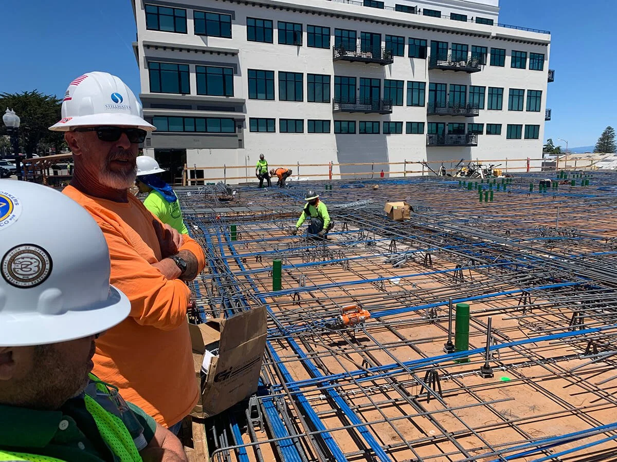 Construction workers on a building site pouring concrete with rebar framework in the foreground, in front of a multi-story building under construction.