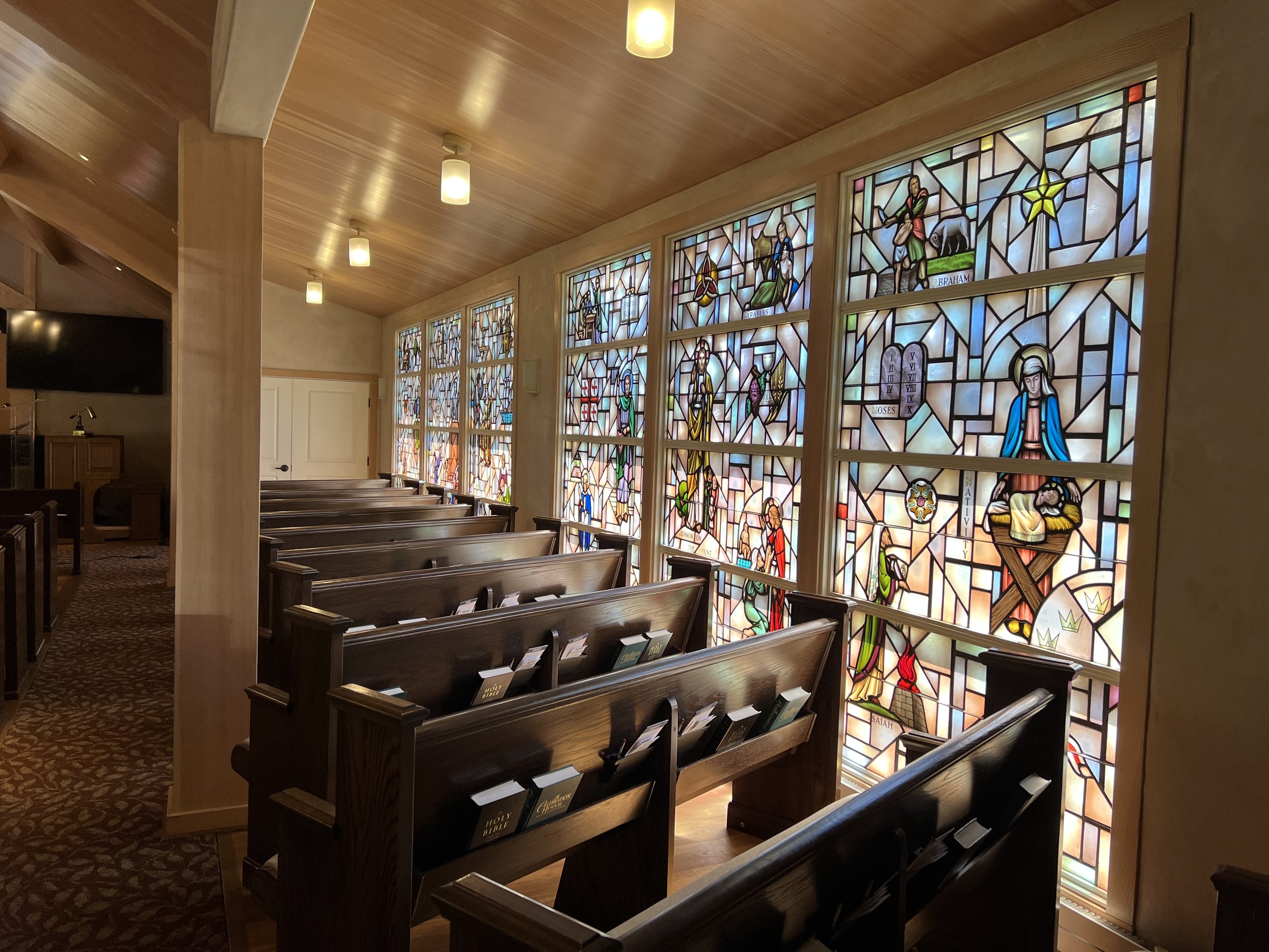 Inside a church with wooden pews and a wall of stained glass windows depicting biblical figures and scenes, illuminated by overhead lights.