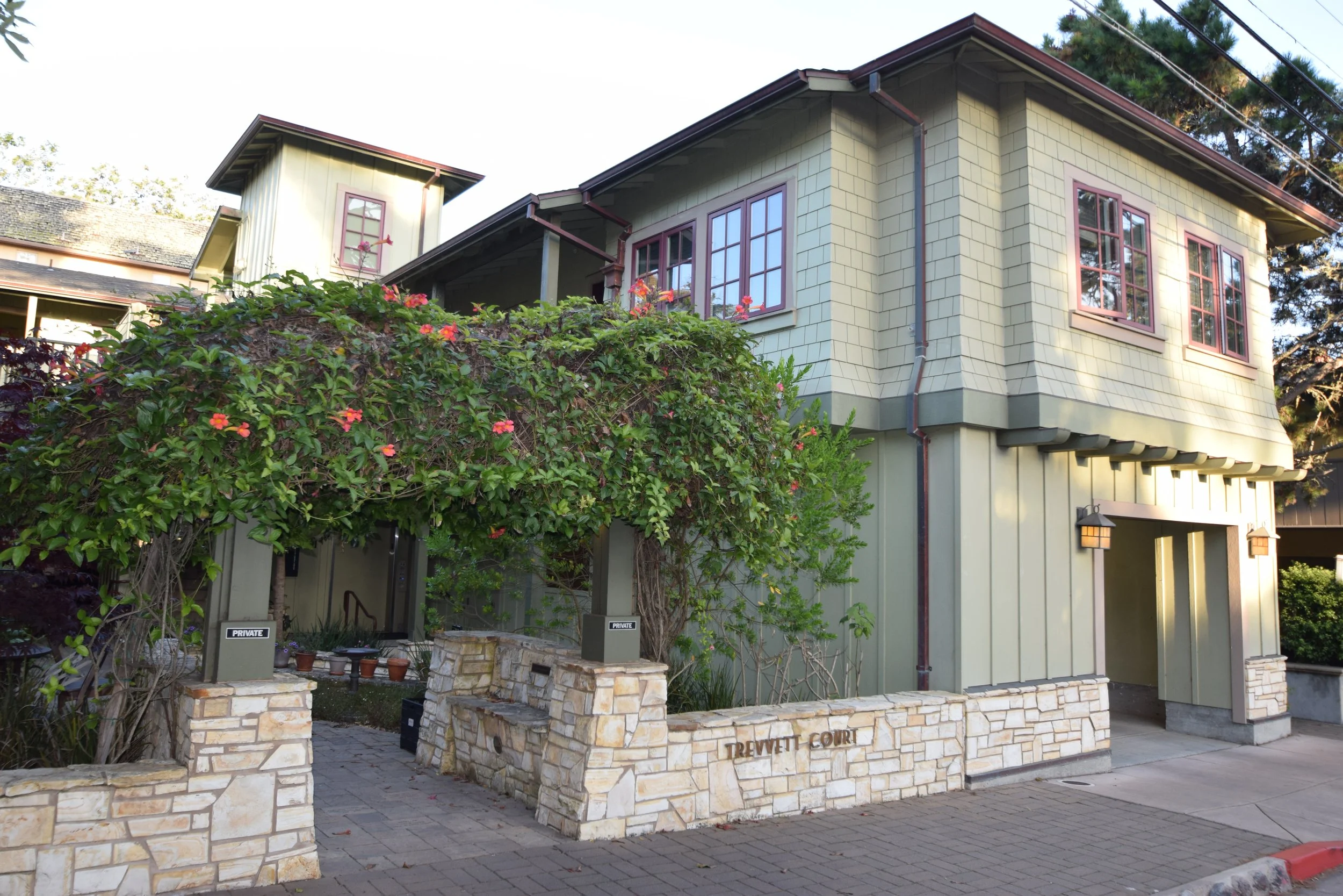 Exterior view of a multi-story beige house with green siding, pink window frames, and stone accents on a low wall with a sign that reads "Trewett Court."