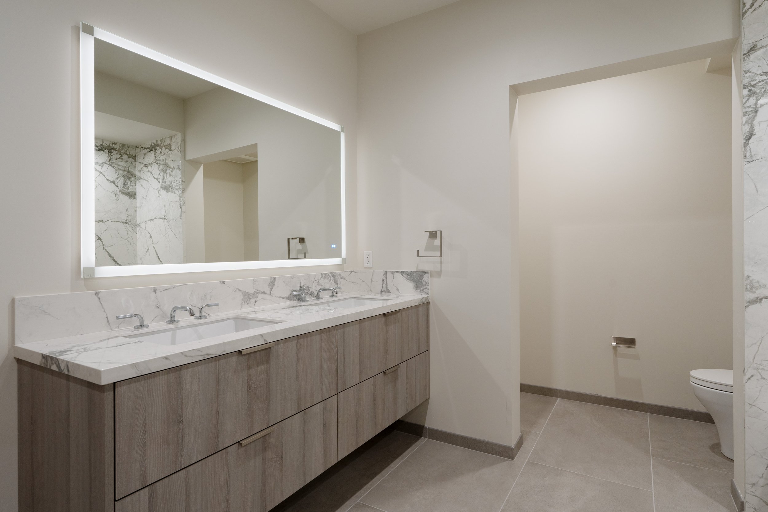 Modern bathroom with double sinks, marble countertops, wooden cabinetry, large illuminated mirror, and toilet, with marble and tile accents.