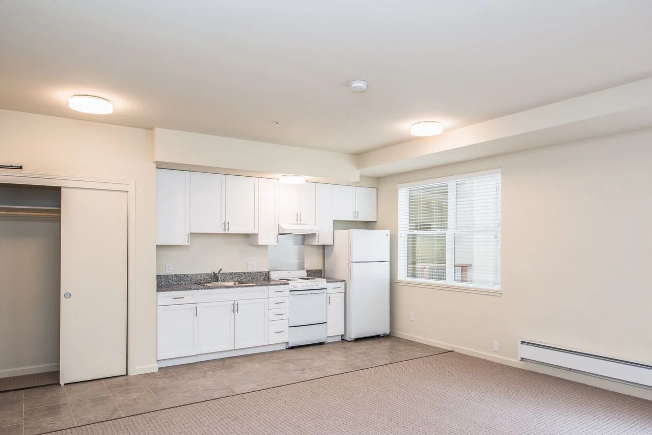Empty apartment with white kitchen cabinets, a refrigerator, a stove, and a window.