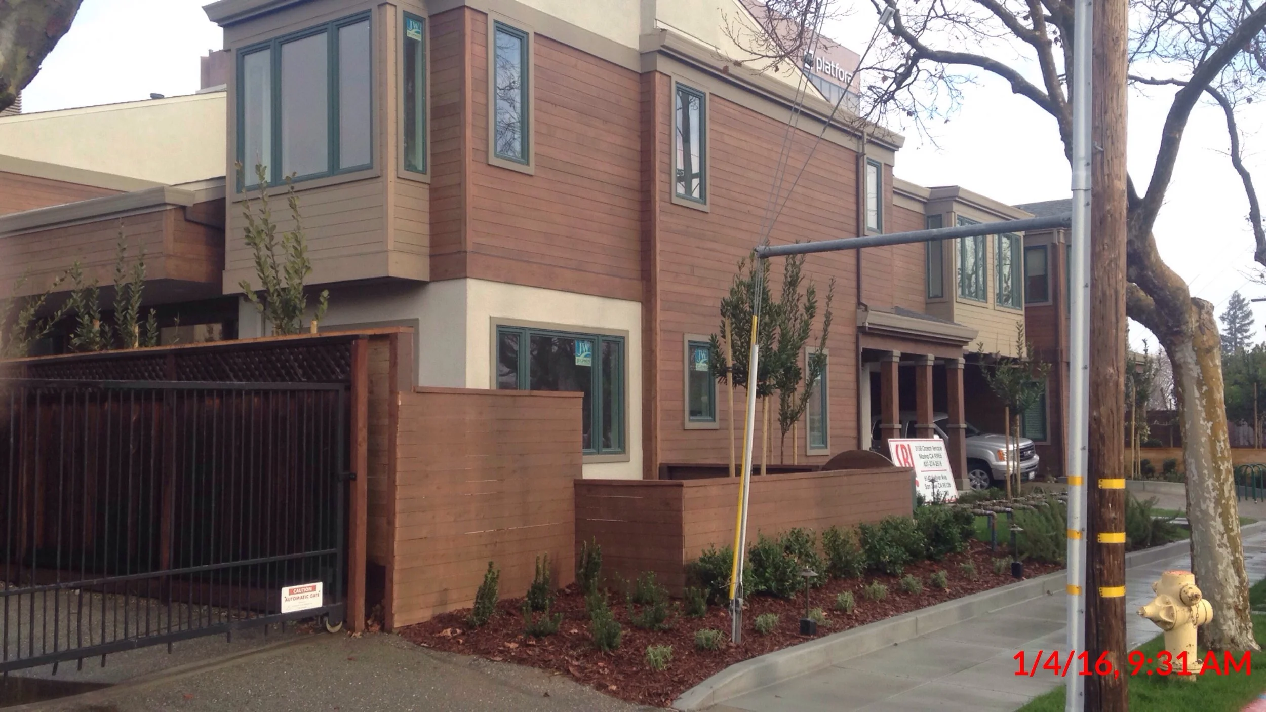 A multi-story residential building with wooden siding and multiple windows. There's a fenced yard with bushes and trees, a street with a sidewalk, and a parked car. Utility poles and a fire hydrant are visible in the foreground.