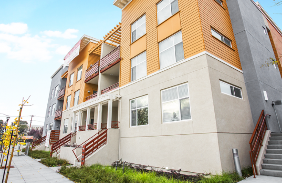 Modern multi-story residential building with beige and orange exterior, multiple windows, and balconies with red railings, set against a blue sky.