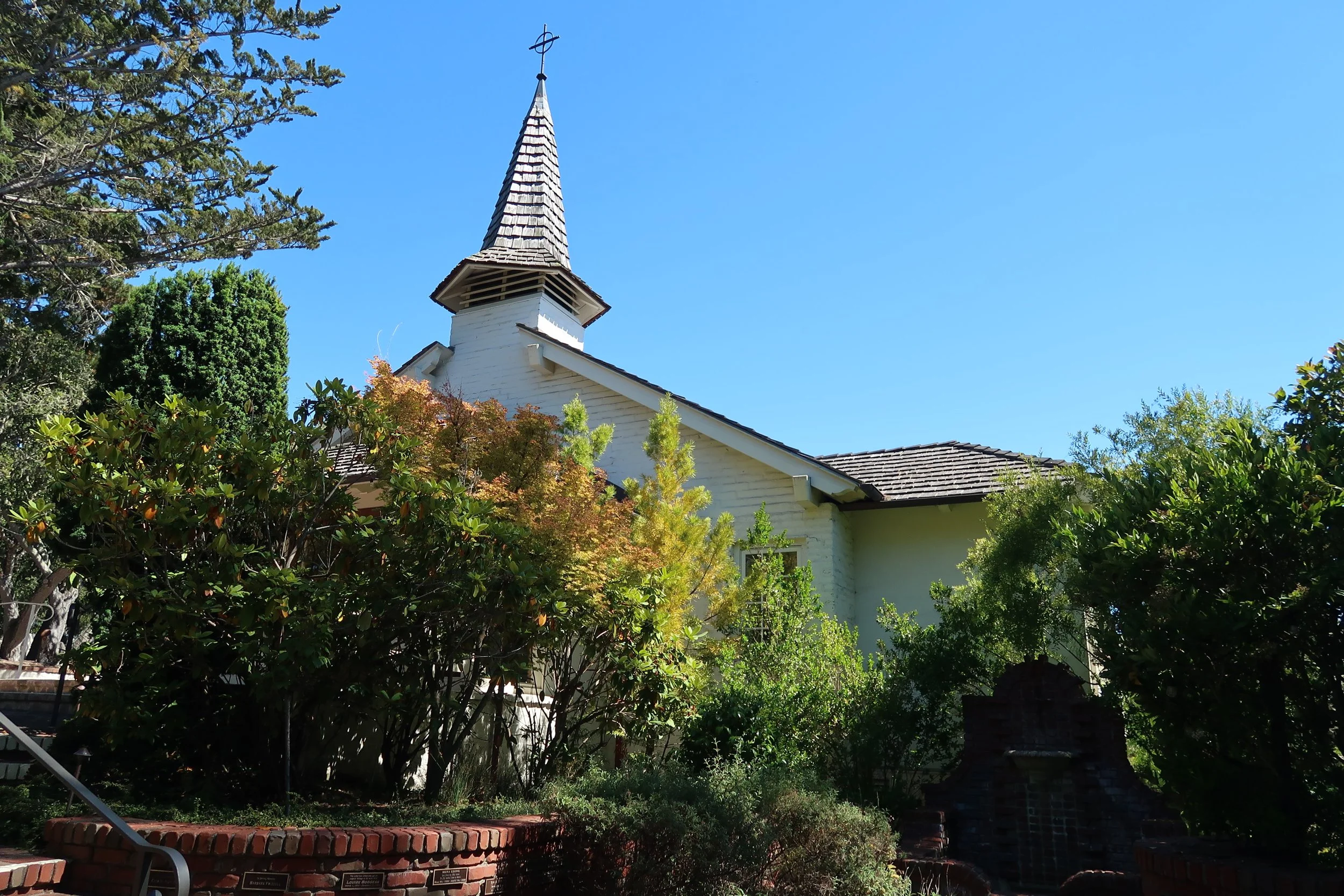 A white church with a tall steeple topped with a cross, surrounded by green trees and bushes, under a clear blue sky.