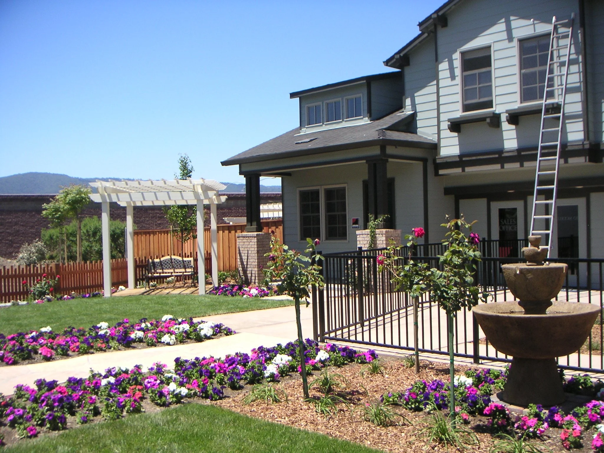 A suburban house with a white exterior, black trim, and a ladder leaning against the second story, surrounded by a landscaped yard with colorful flowers, a fenced patio area, and a white pergola.