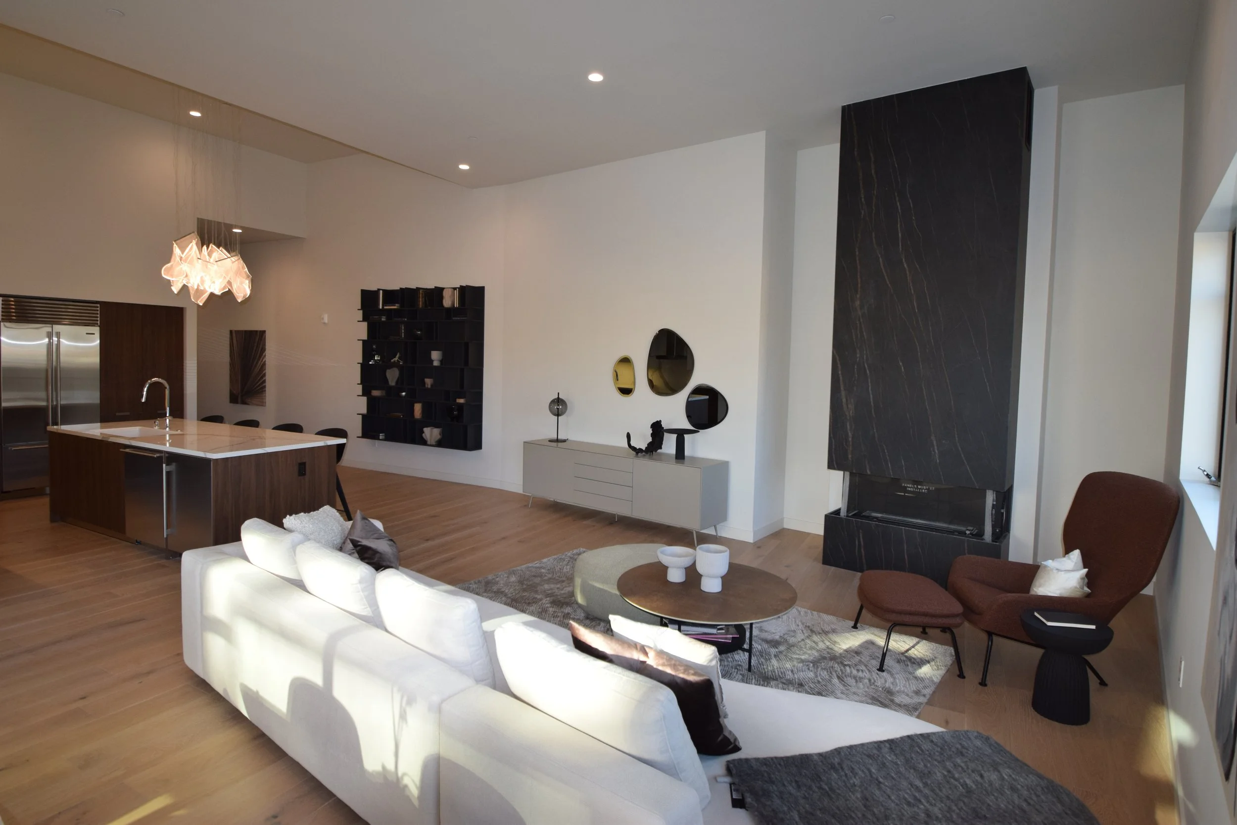 Modern living room with white sofa, two brown armchairs, a coffee table, and a black fireplace with gold veins. There's a white sideboard with decorative items and a black bookshelf with books. The room has wooden floors and recessed ceiling lights.