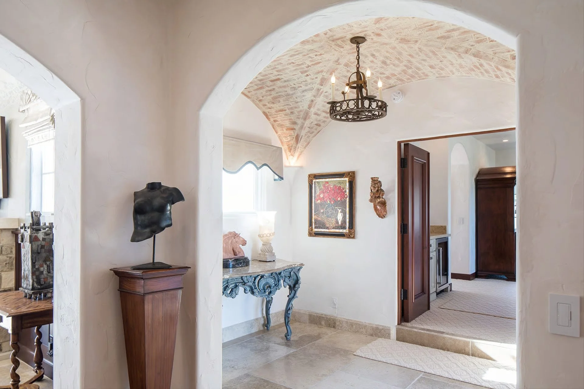 Interior of a house with a vaulted brick ceiling, a chandelier, and art on the walls. A small sculpture of a torso on a pedestal is visible, along with a decorative table and a framed picture.