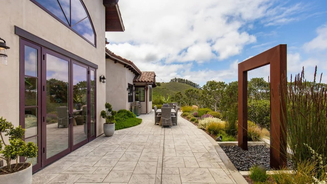 Outdoor patio area with dining table, chairs, and plants, adjacent to a modern house with large glass doors and a scenic view of hills and greenery under a partly cloudy sky.