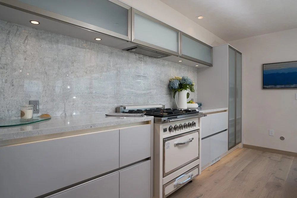 Modern kitchen with white cabinets, a marble backsplash, and a stove with a sink on the right side. There is a vase with blue and green flowers on the counter. A flat-screen TV is mounted on the wall to the right.