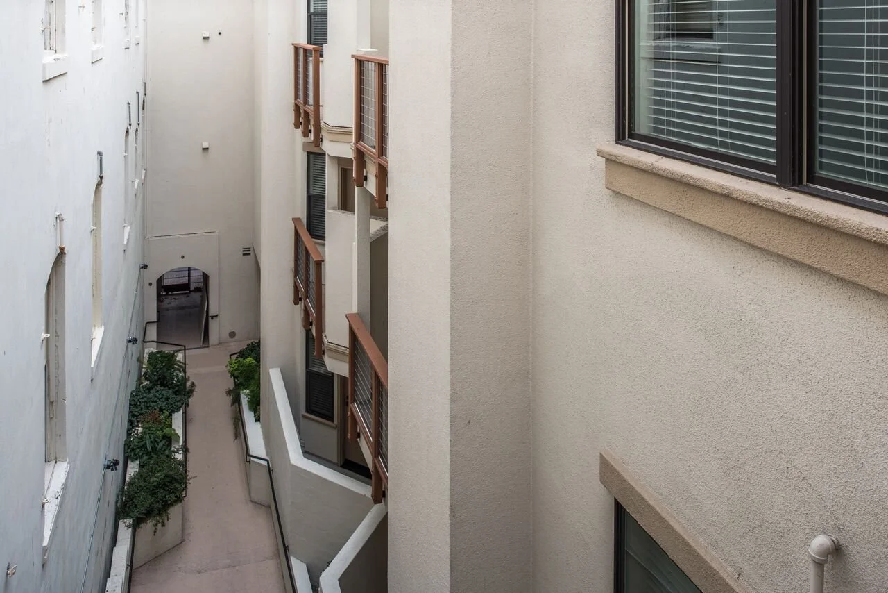 View of an alley between two beige apartment buildings with windows, some with blinds, small balconies with brown railings, potted plants, and a pathway leading to an arched doorway at the back.