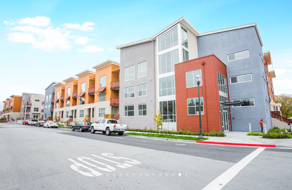 A modern apartment complex with multiple stories, colorful exterior walls, and parking in front, with several parked cars and a person walking on the sidewalk.