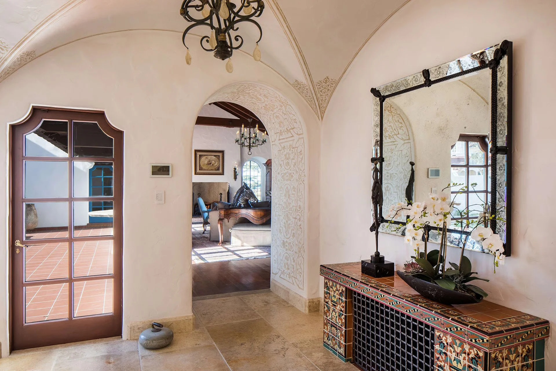 View of an elegant entrance hallway with a large mirror, a colorful tiled console table, and decorative arches. A chandelier hangs from the ceiling, and a glass door leads to an outdoor patio with terracotta tiles and potted plant, leading into a liv