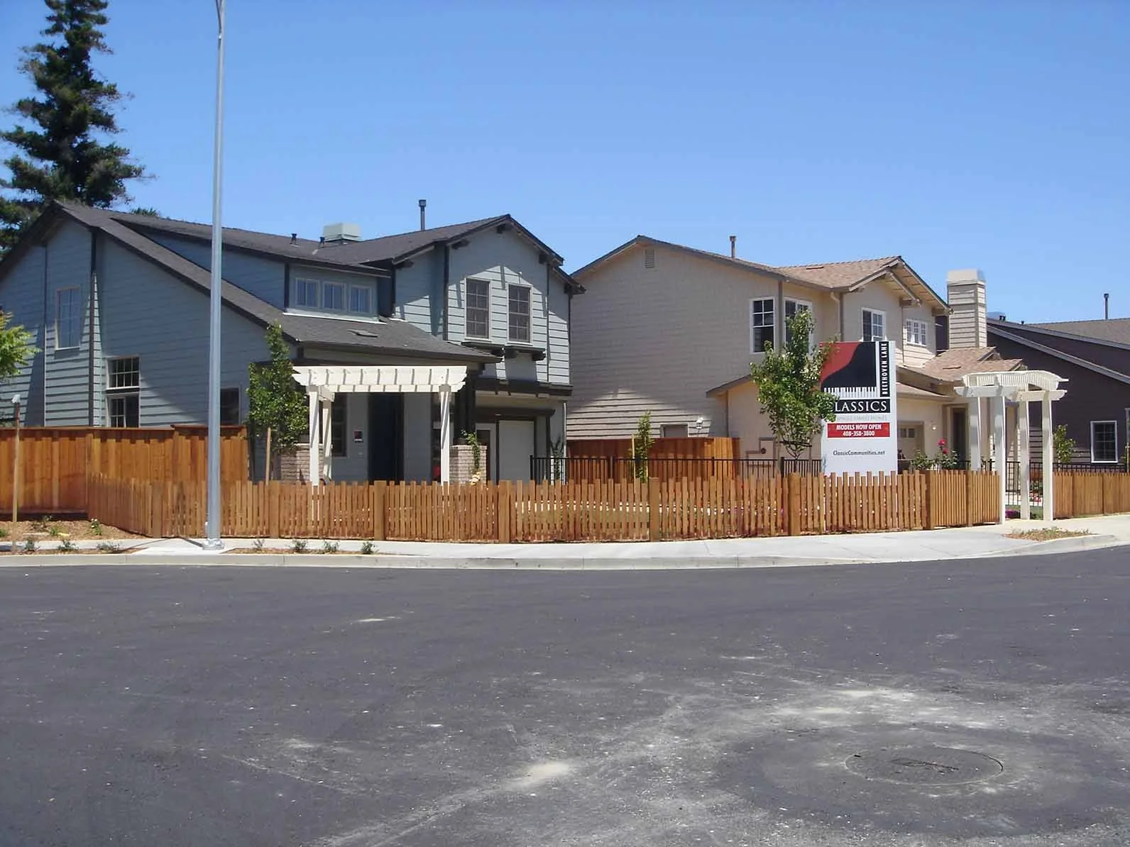 Two modern houses with wooden fences and a sign for classic models, with a clear blue sky and a sidewalk in front.