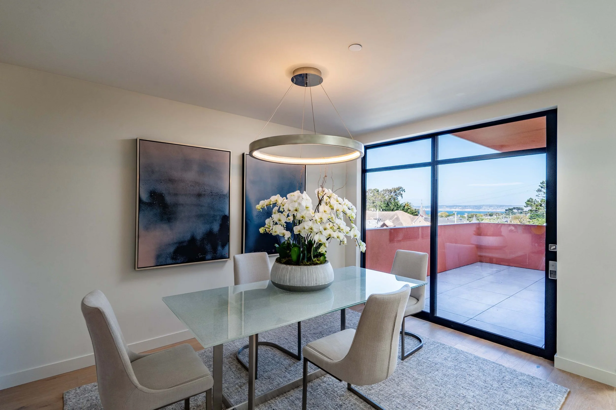 Modern dining room with a glass table, six beige upholstered chairs, a large white vase with white orchids, abstract wall art, a ceiling light fixture, and large floor-to-ceiling windows showing a view of a balcony and the outside landscape.