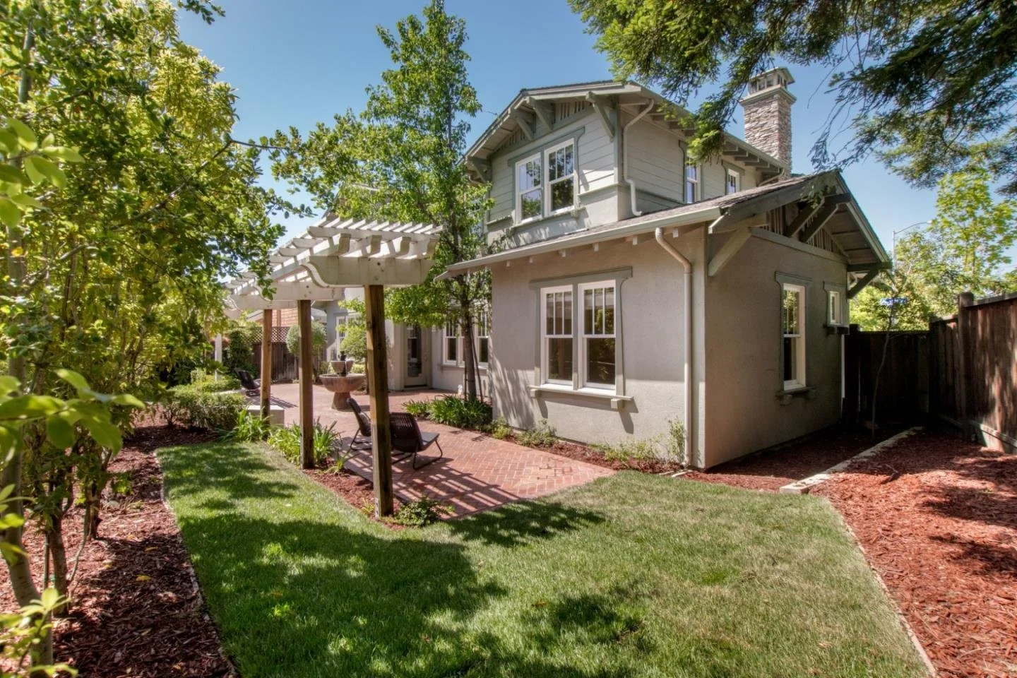 A backyard with a small lawn, brick patio, and outdoor seating under a pergola, with a two-story house and trees in the background.