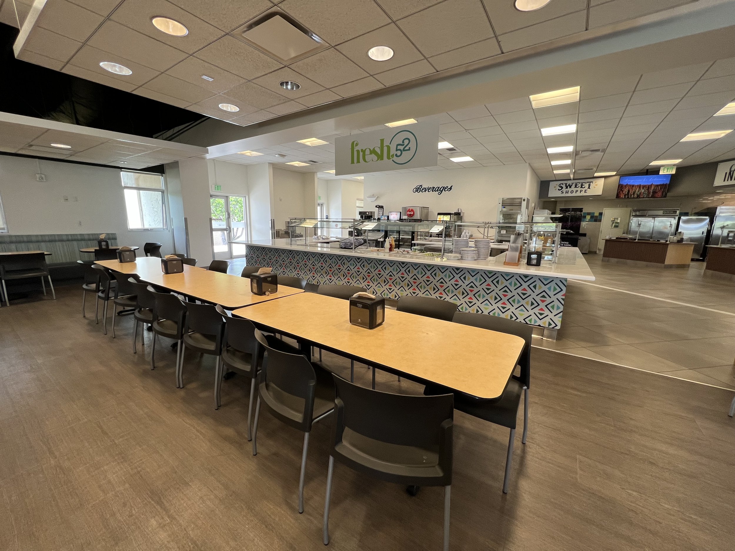 Interior of a casual dining area with a long wooden table, black chairs, and a counter area with a geometric patterned front. There is a sign that reads 'fresh 52' and various beverage machines. The space is well-lit with ceiling lights and has large