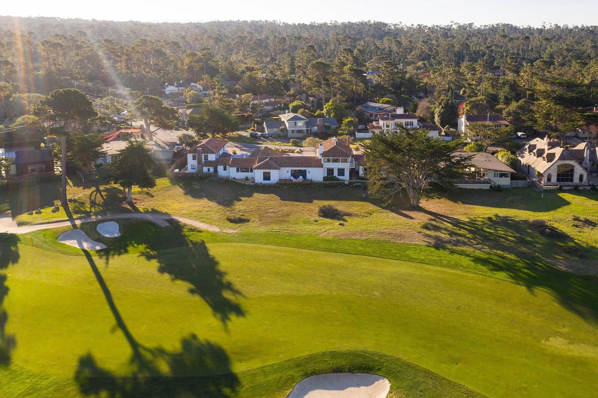 An aerial view of a residential neighborhood with houses, trees, and a golf course with sand bunkers in the foreground, during daylight with shadows cast by trees.