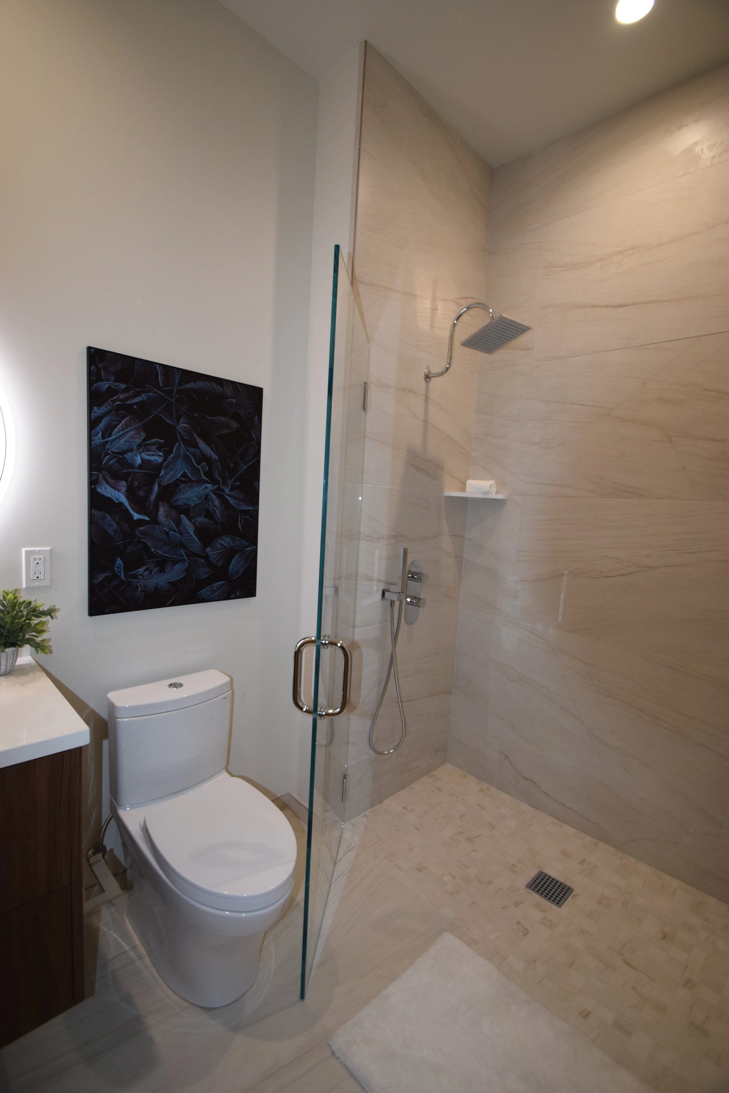Modern bathroom with a walk-in shower, beige marble walls, a wall-mounted showerhead, a toilet, a small cabinet with a plant, a piece of wall art, and a white bath mat.