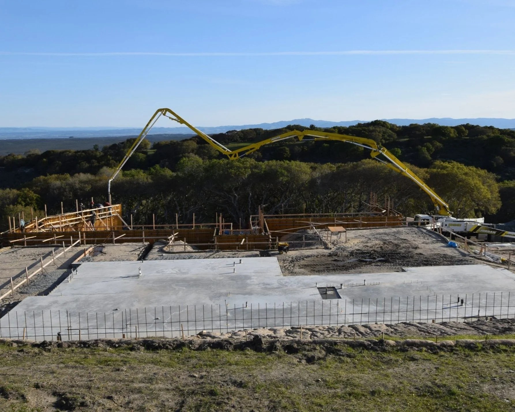 Construction site with concrete foundation, wooden forms, and a large yellow concrete pump truck working on a hilltop with trees and distant mountains in the background.