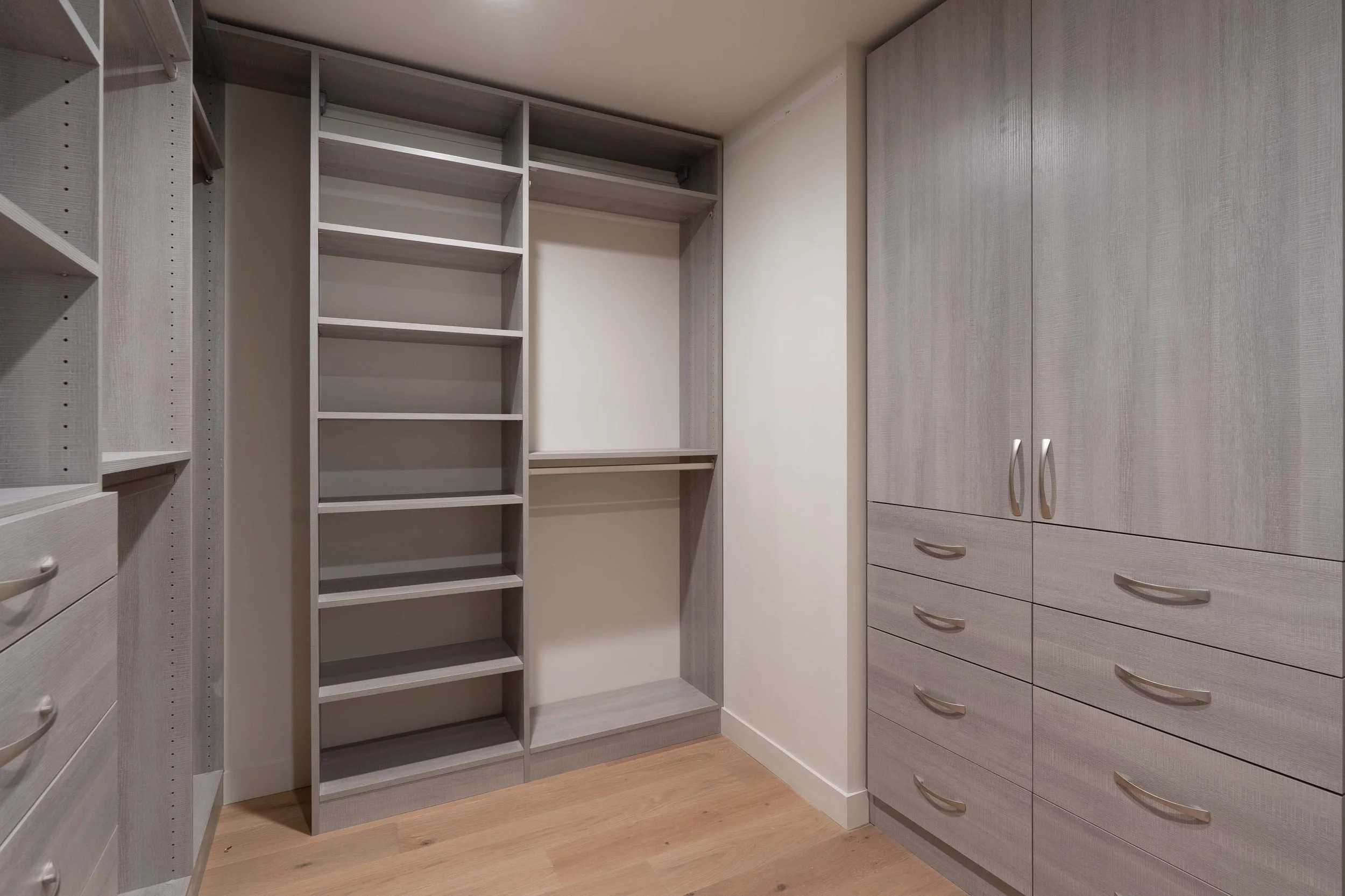 Empty walk-in closet with built-in shelves, drawers, and a hanging rod, with light-colored wood cabinetry and flooring.