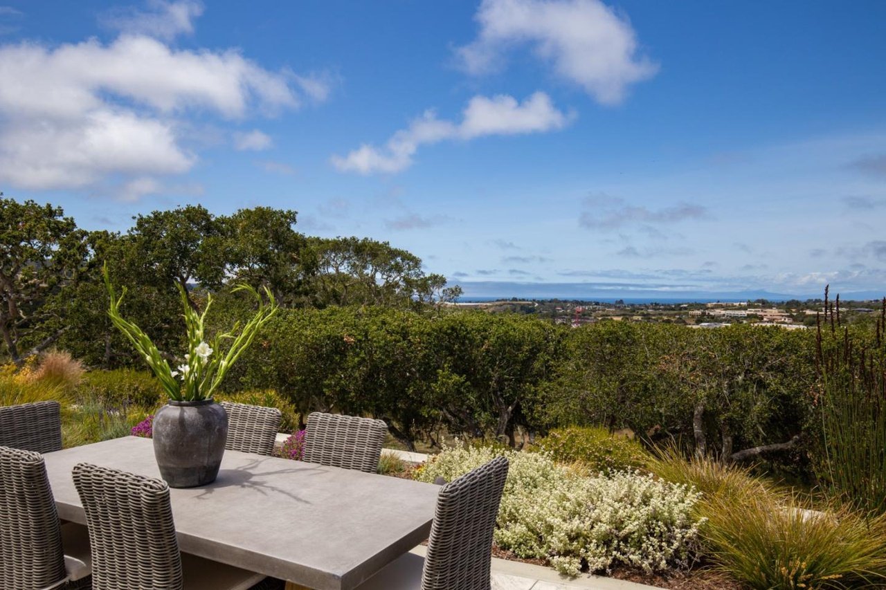 Outdoor patio with a table and wicker chairs, a large vase with white flowers, surrounded by greenery, with a view of trees, a cityscape, and the ocean in the distance under a partly cloudy sky.