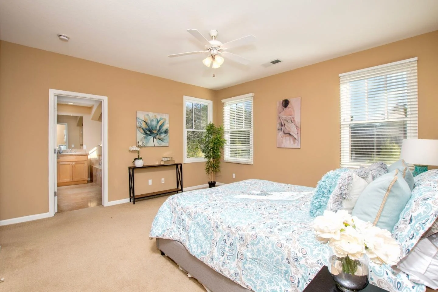A bedroom with tan walls, a bed with blue and white bedding, multiple pillows, and a bouquet of white flowers on a nightstand. Two windows with white blinds, a ceiling fan, artwork on the walls, and an open doorway showing a bathroom with a wooden va
