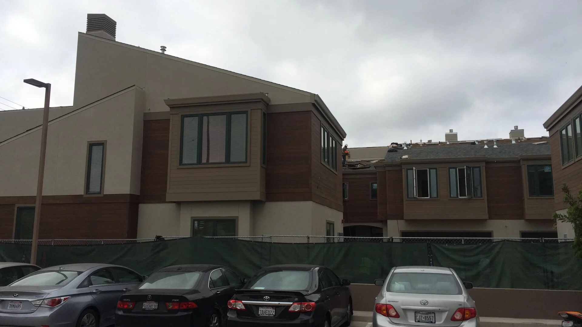 Multi-story residential buildings under construction, with workers on the roof, a parking lot with several cars in the foreground, and an overcast sky.