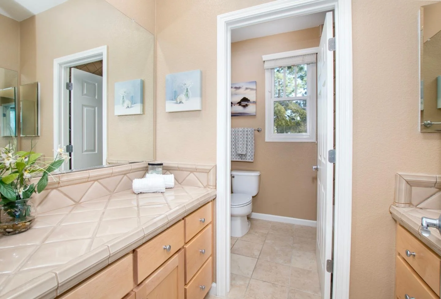 Bathroom with beige walls, a large mirror above a beige tiled counter, a small towel, a vase with flowers, a window over the toilet, and framed artwork on the walls.