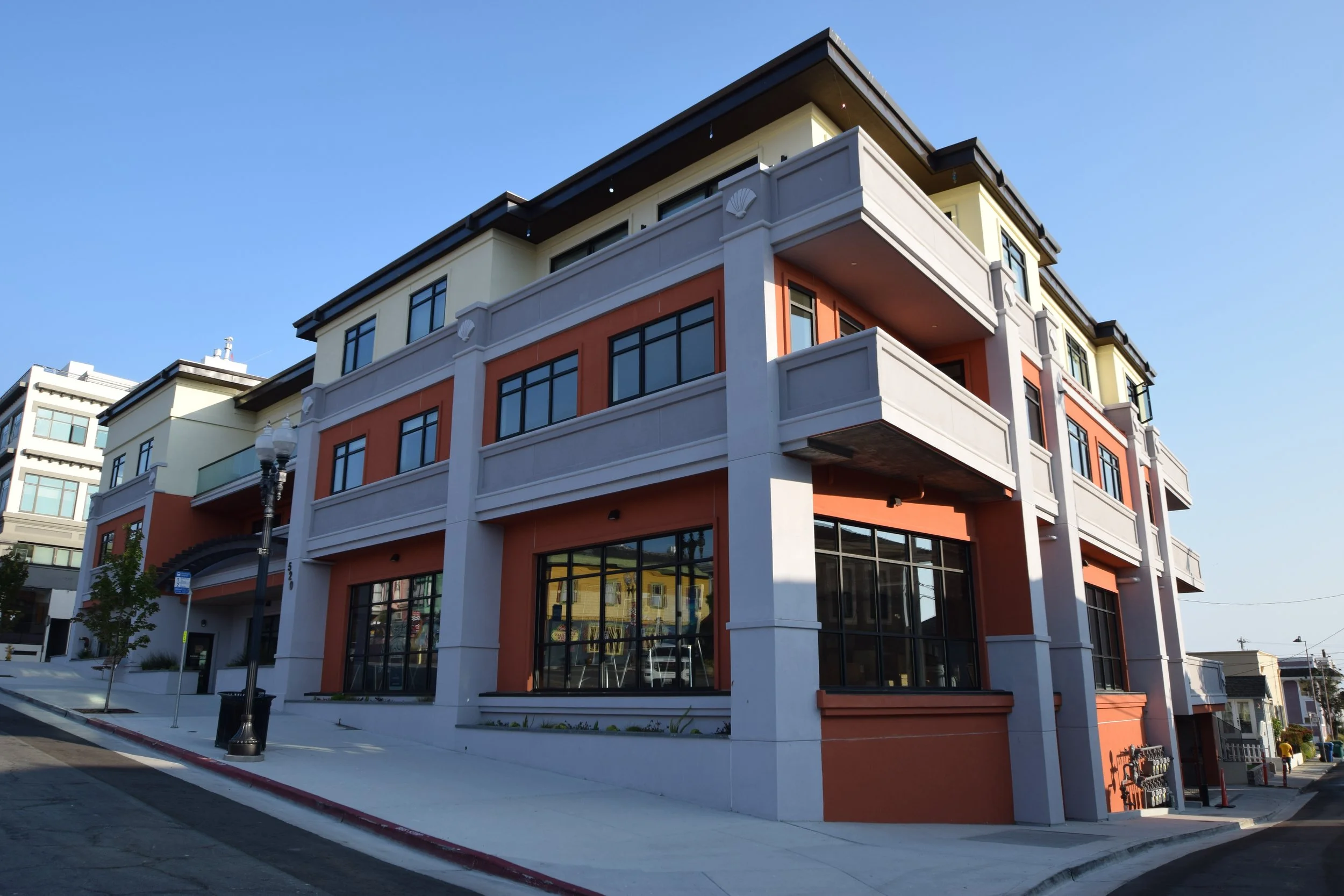 Modern multi-story residential building with large windows, balconies, and colorful exterior walls in shades of white, orange, and yellow. The building is situated on a street corner with a sidewalk, street lamps, and a clear blue sky overhead.