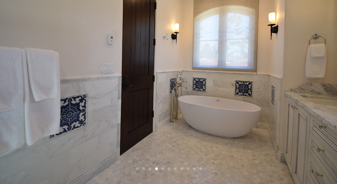 Bathroom with a freestanding bathtub, marble tile accents, two wall sconces, a large window with a blind, a towel on the left, and a vanity with a marble countertop on the right.